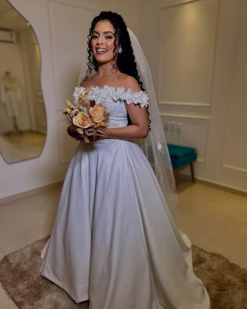 Modelo de noiva com cabelos cacheados pretos e compridos, com maquiagem e penteado de cerimônia, véu, vestido branco encorpado com flores brancas formando as alças e um buquê de flores secas