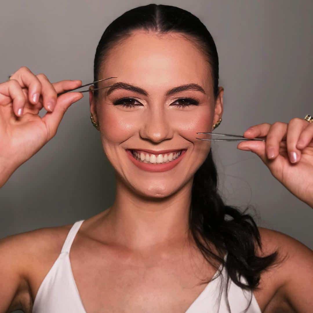 Madu Damasceno, designer de sobrancelhas em Governador Valadares no Ateliê SF Beauty, maquiada, sorrindo e segurando duas pinças