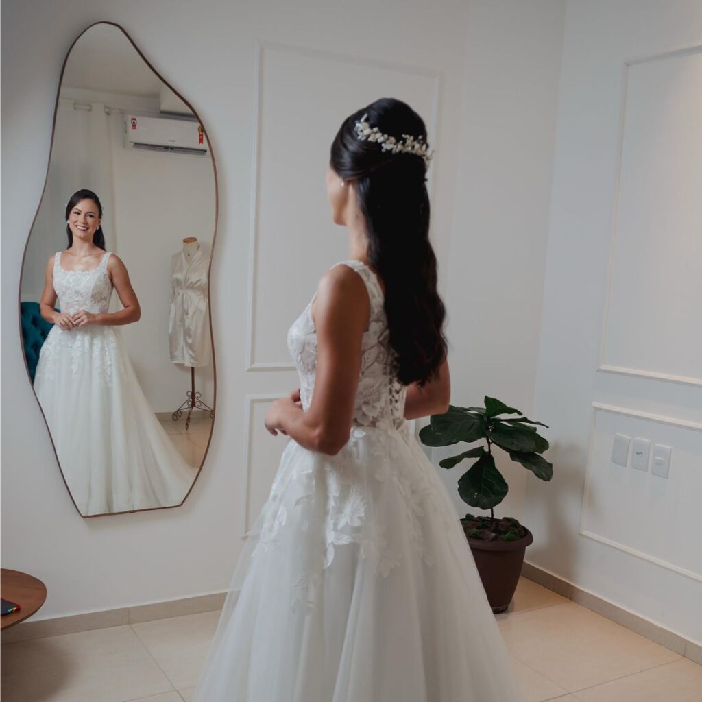Noiva refletida no espelho, sorrindo e produzida no Ateliê SF Beauty em Governador Valadares, com maquiagem beauty resistente, penteado semipreso com grinalda, cabelos negros ondulados e vestido de noiva elegante