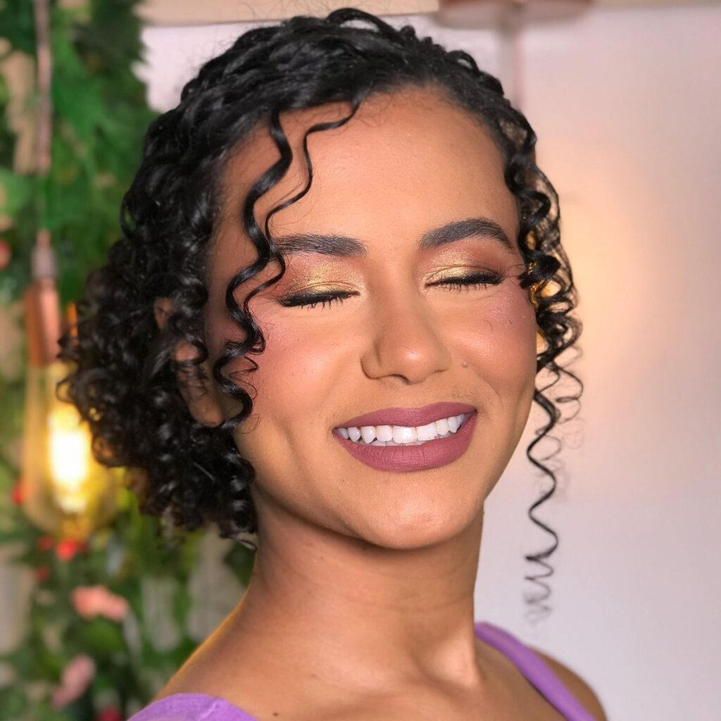 Maquiagem em Governador Valadares delicada e elegante com sombras nos olhos indo do dourado no centro migrando para um castanho avermelhado nas extremidades, com brilho suave. Pele morena clara, penteado de cabelos cacheados com coque delicado e sutil. Sobrancelhas bem feitas. Sorrindo com batom matte cor malva