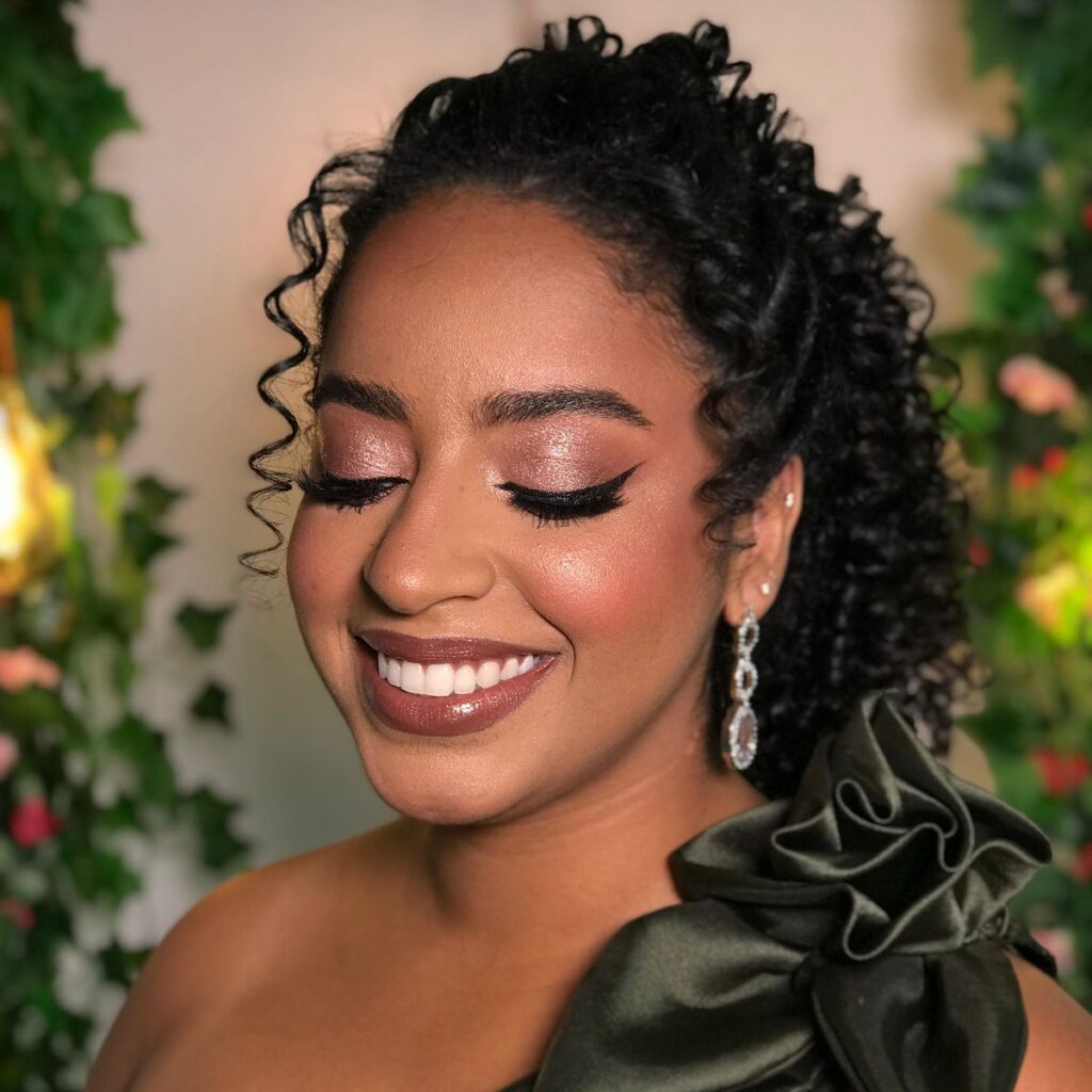 Maquiagem suave em Governador Valadares, com sombras sutis, brilho nas pálpebras e delineado gatinho. Penteado semipreso de cabelo cacheado, muito bem elaborado, batom castanho nude com gloss brilhante e um belo sorriso. Pele morena clara.